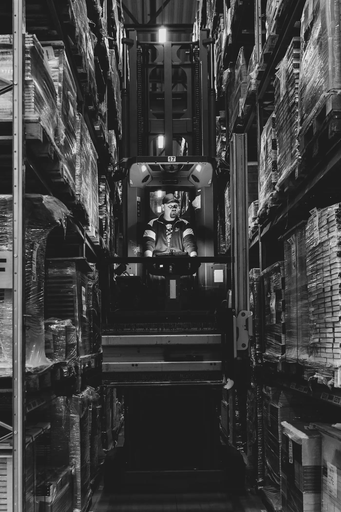An operator driving a high-reach truck down a narrow aisle between tall pallet racks in a distribution warehouse.